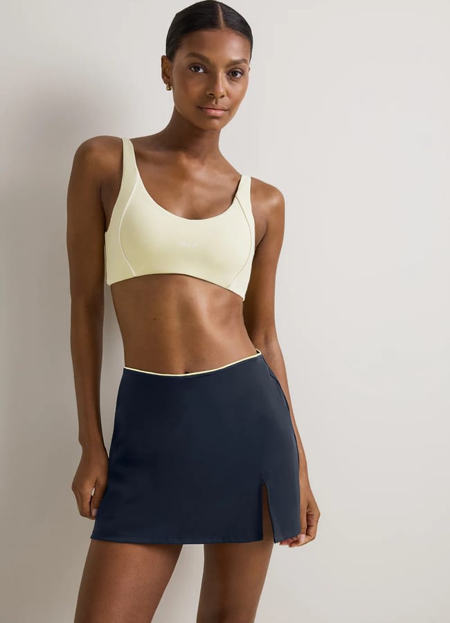 A woman stands against a plain light-coloured wall, wearing a pale yellow sports bra and a high-waisted navy blue athletic skirt with a small side slit. She looks forward with a relaxed expression, perfectly capturing modern court fashion.