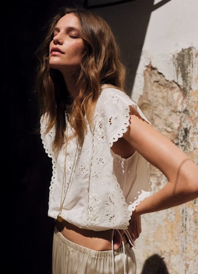 A woman with wavy brown hair stands in sunlight, eyes closed, wearing a sleeveless white broderie anglaise summer top and light-coloured trousers, with a long necklace. She is next to a rough, textured wall.