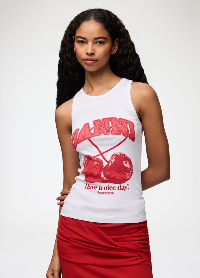 A woman with long, wavy hair wears a sleeveless white top featuring fruit prints and the text “GANNI Have a nice day!” She pairs it with a red skirt and stands against a plain, light background.