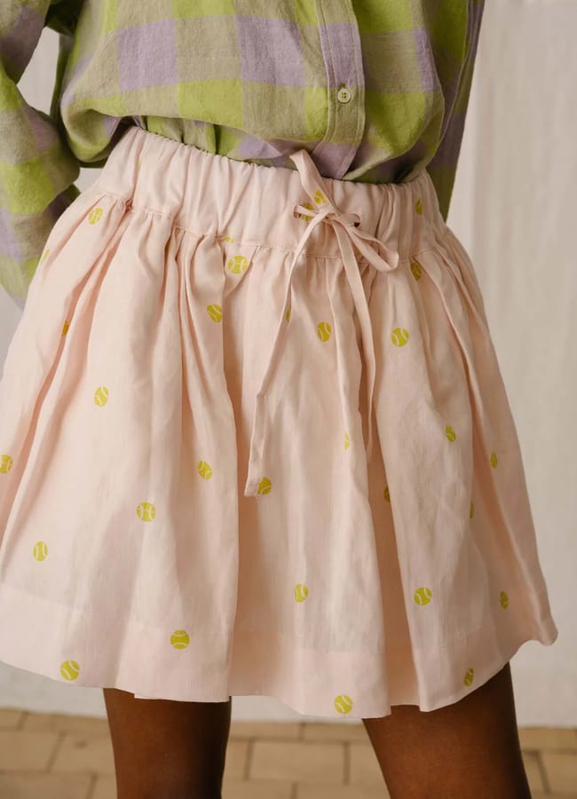 A person wearing a pastel green and purple checked shirt tucked into a light pink skirt with an elastic waistband, bow tie, and yellow tennis ball pattern captures the playful spirit of cute padel outfits. Only the lower torso and upper legs are visible.