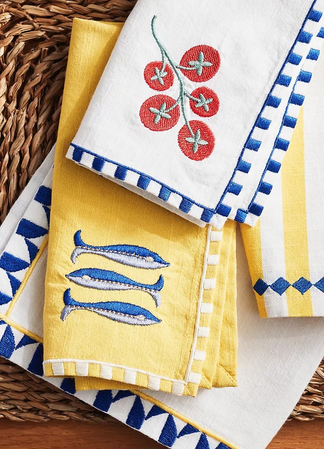 A close-up of colourful embroidered serviettes: one white with red tomatoes, one yellow with blue fish, and striped or chequerboard blue and yellow borders, arranged on a woven mat.
