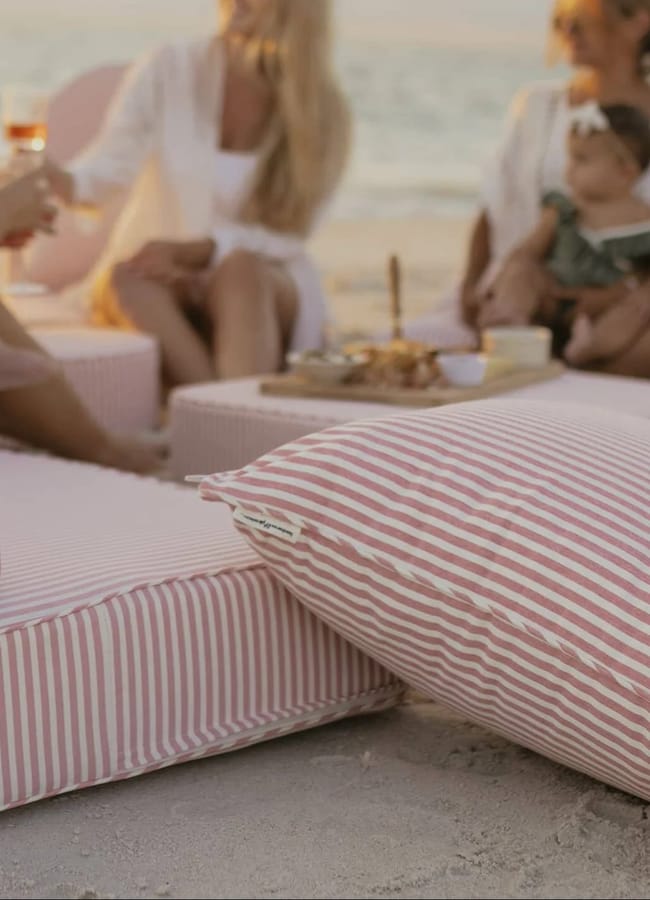 Women and a baby sit on pink and white striped cushions at the beach, relaxing near a low table with picnic essentials and snacks. The scene is casual and serene, set near the sea.
