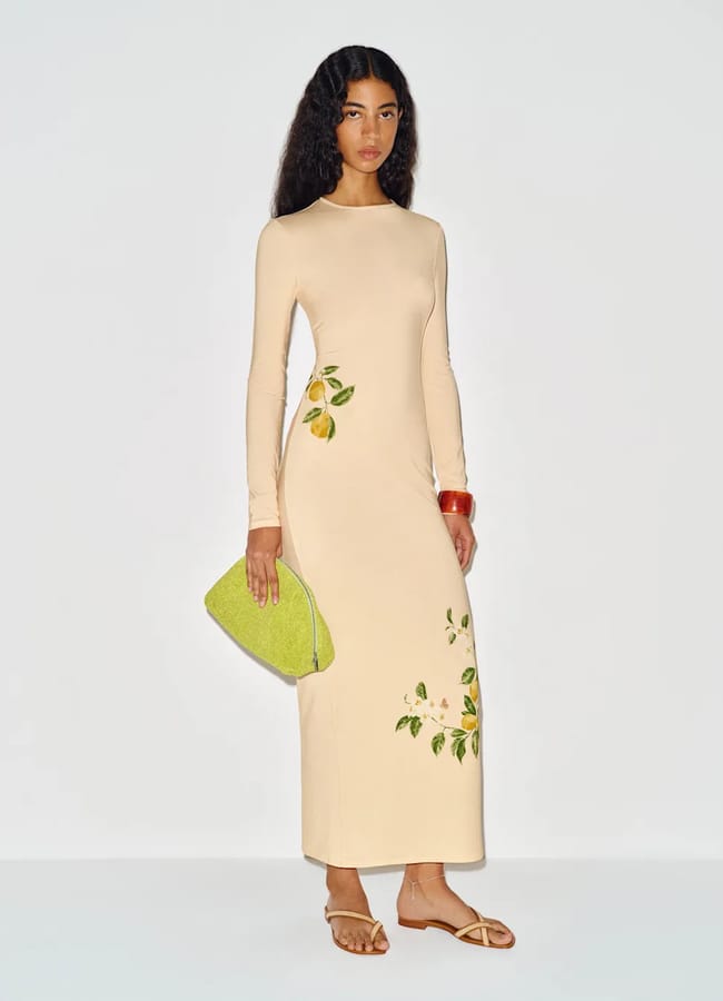 A woman with long wavy hair stands against a plain background, wearing a long-sleeved, light beige dress adorned with fruit prints of painted lemons and leaves. She holds a large green clutch, pairing it with tan sandals and a chunky bracelet.