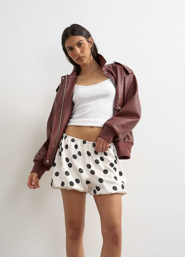 A woman stands against a white wall wearing a maroon leather jacket, a white crop top, and playful bubble shorts with large black polka dots. She looks at the camera with a relaxed expression and one hand in her pocket.