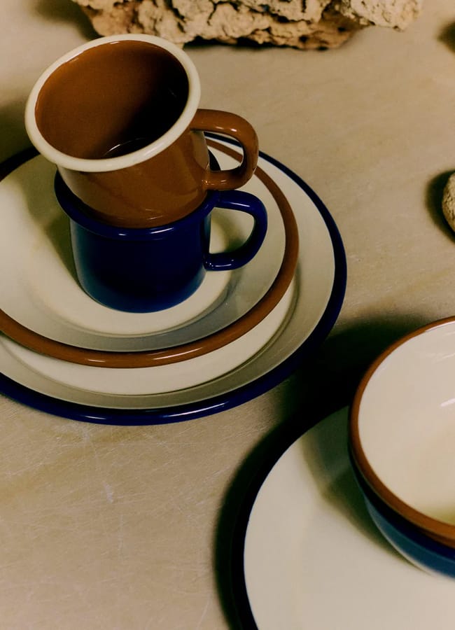 Two stacked plates and two stacked cups, in brown, cream, and blue colours, sit on a beige surface next to a matching bowl. These modern, minimalist pieces are picnic essentials for stylish outdoor dining.