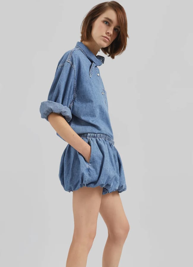 A person with short brown hair wears a blue denim long-sleeved shirt with rolled sleeves and matching loose-fitting bubble shorts, standing against a plain light grey background.