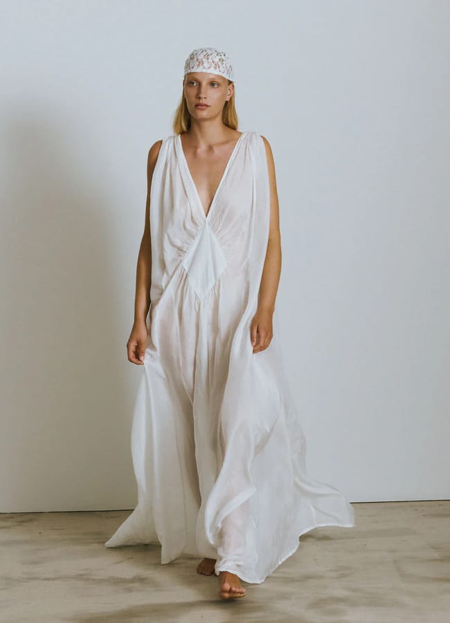 A woman wearing a flowing, sheer white gown with a deep V-neckline walks barefoot on a concrete floor—her look is serene, almost ethereal, perfect inspiration for unique Fathers’ Day gifts 2026. She has straight blonde hair and wears a decorative white headpiece.