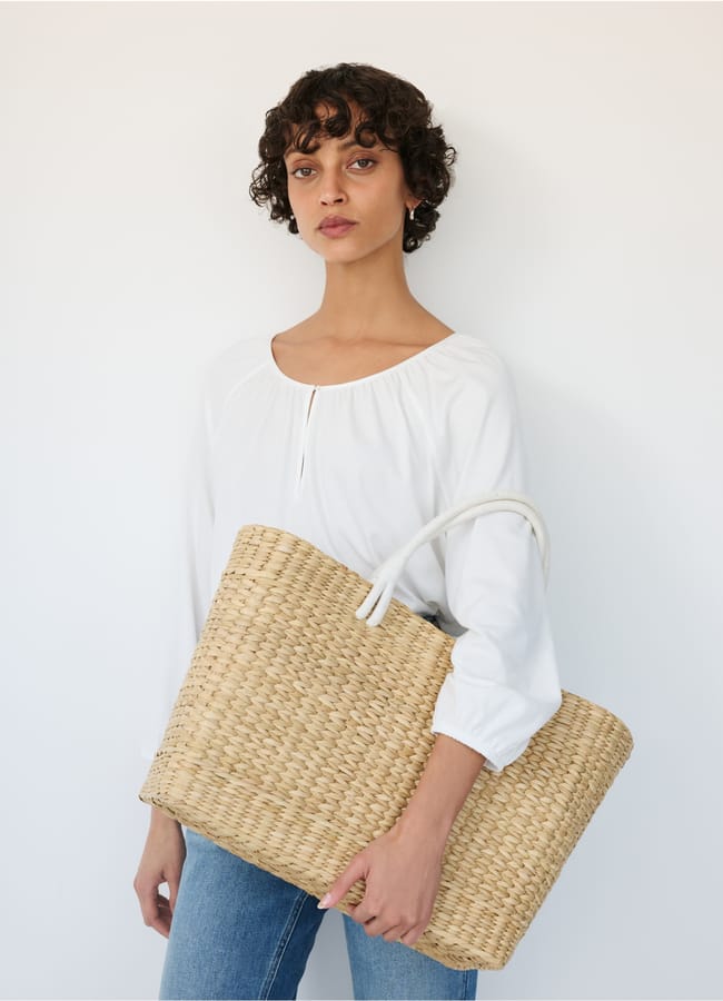 A woman with short curly hair, wearing a white long-sleeved blouse and blue jeans, stands against a plain white background holding a large woven straw basket bag.