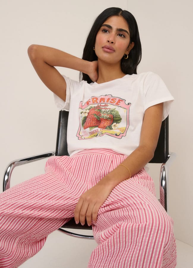 A woman sits on a black chair, wearing a white T-shirt with a strawberry fruit print and the word Fraise, paired with pink and white striped trousers. She has long dark hair and rests one arm behind her head.