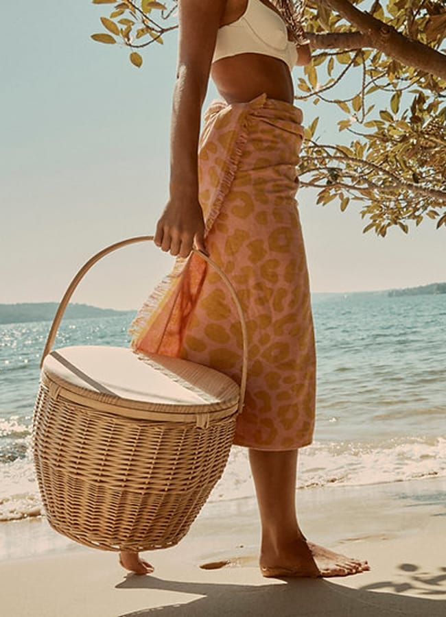 A person in a white bikini top and pink animal-print sarong stands barefoot on a sandy beach, holding a woven basket filled with picnic essentials, with water and tree branches in the background.