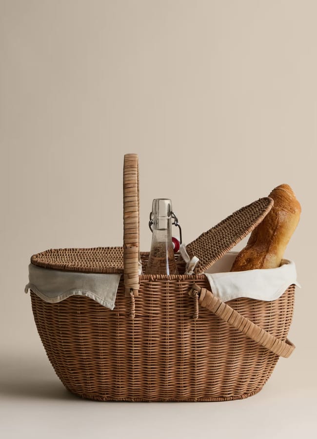 A wicker picnic hamper with a cloth lining, filled with picnic essentials—a bottle, a baguette, and a small red item—sits against a plain beige background. The hamper’s lid is partially open.