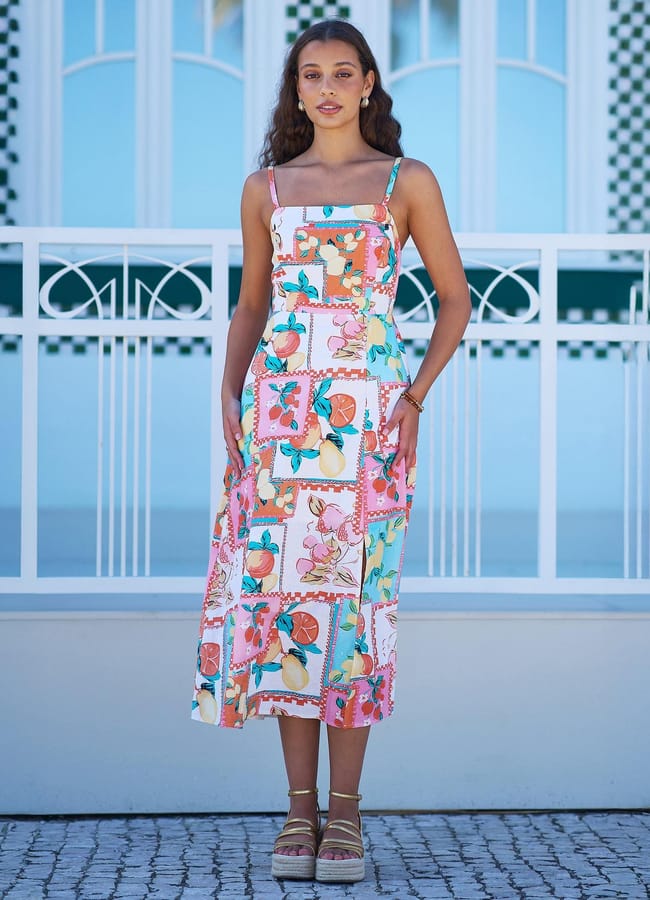 A woman stands outdoors wearing a colourful patchwork sundress with fruit prints and spaghetti straps, paired with wedge sandals; she poses in front of a blue and white building with decorative railings and tiled pavement.