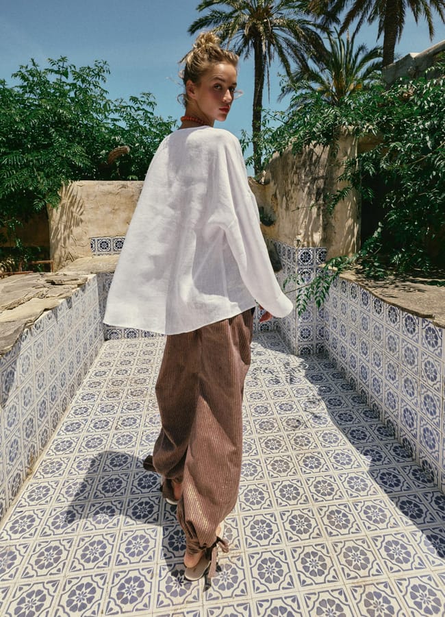 A woman wearing a loose high street linen white top and brown wide-leg trousers stands on a patterned tile floor in an outdoor setting with greenery and palm trees under a clear blue sky.