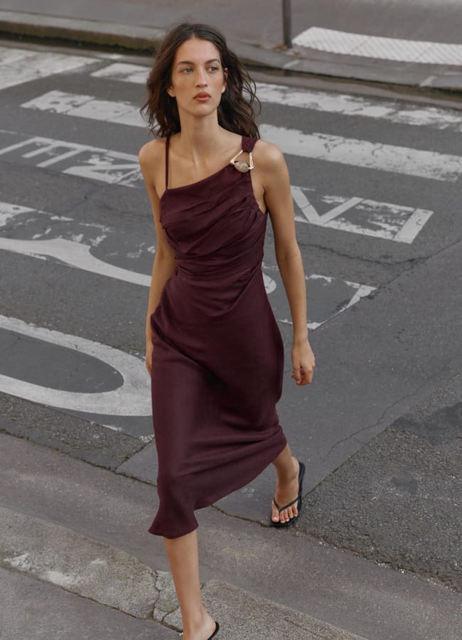 A woman wearing a sleeveless, dark burgundy high street linen dress with an asymmetric neckline and black sandals walks across a street with a faded zebra crossing.