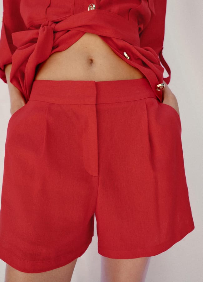 A person wearing a red high street linen outfit with a tied shirt exposing their midriff and matching high-waisted shorts, hands in the pockets, standing against a neutral background.