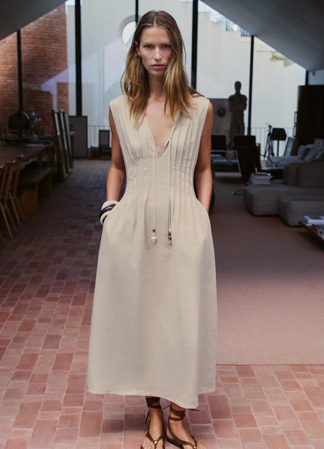 A woman with long hair stands indoors on a brick floor, wearing a high street linen sleeveless, light beige midi dress with a deep V-neck and drawstrings, paired with brown sandals. The room features modern furniture and soft natural light.