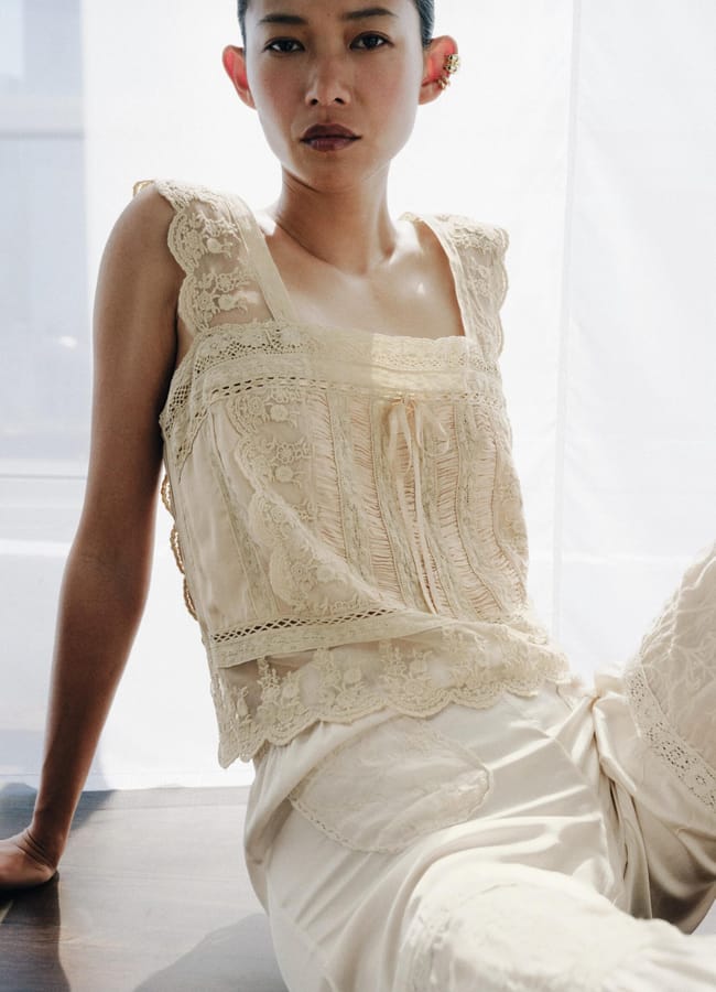 A person with short dark hair sits by a window, wearing an elegant, sleeveless cream-coloured lace summer top and matching trousers. The soft lighting highlights the delicate details of the outfit.