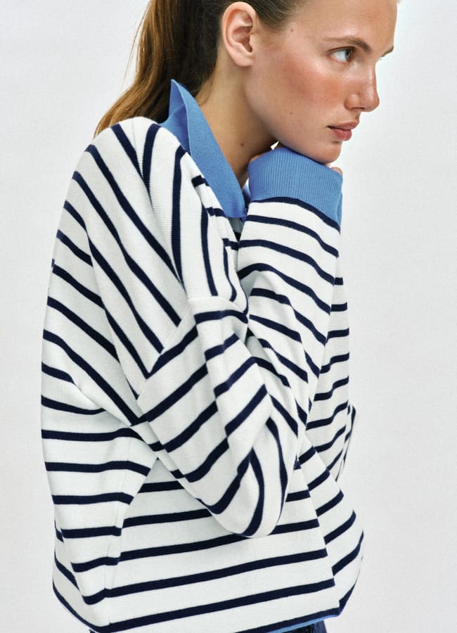 A woman with light brown hair in a ponytail wears a white jumper with navy blue stripes over a blue collared shirt—a chic nod to court fashion. She stands in profile, looking right, hands near her face.