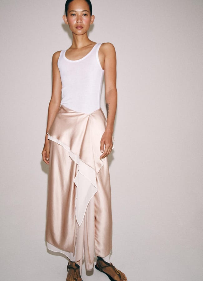 A person stands against a plain background wearing a white sleeveless vest, a light pink satin wrap skirt with layered frills, and brown flat sandals—a minimalist, elegant look perfect for Father's Day gifts 2026 inspiration.