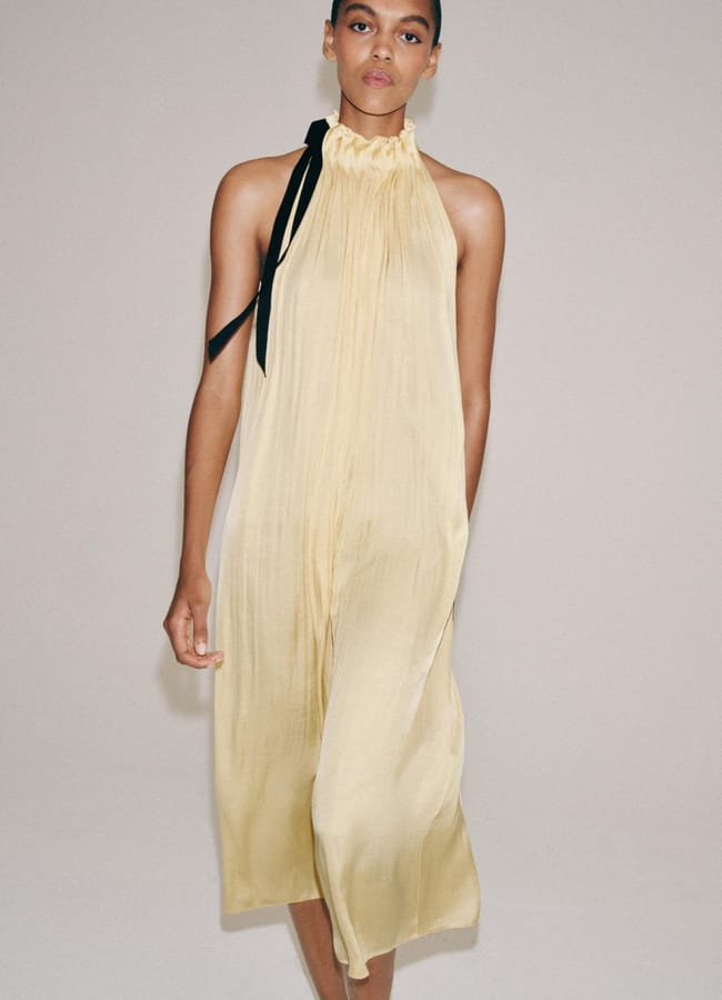 A woman wearing a sleeveless, pale yellow, pleated dress with a high ruffled neckline and a black ribbon tied at the shoulder stands against a plain light background, offering fresh inspiration for Fathers’ Day gifts 2026.