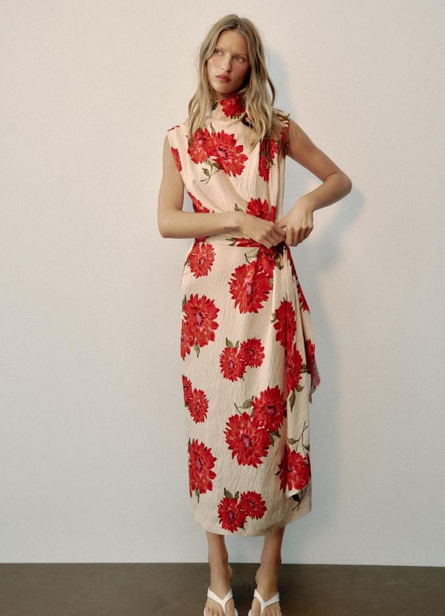 A woman wearing a sleeveless, ankle-length cream dress with large red floral prints—a beautiful example of high street fashion—stands against a plain wall. She has loose, wavy hair and is wearing white flip-flops.