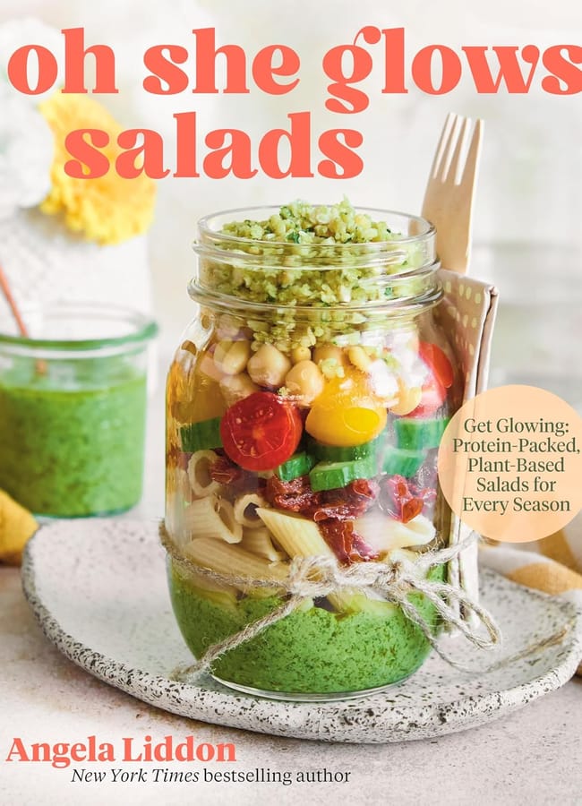 A cookbook cover titled oh she glows salads by Angela Liddon, featuring a kilner jar layered with pasta, cherry tomatoes, chickpeas, greens, and pesto—perfect inspiration for salad season—set on a light-coloured surface.