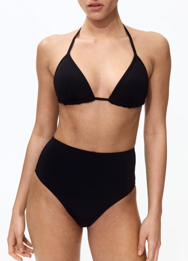 A woman models a black bikini with a halter-style triangle top and high-waisted bottoms against a plain white background—an effortlessly chic option for what to wear in Ibiza.