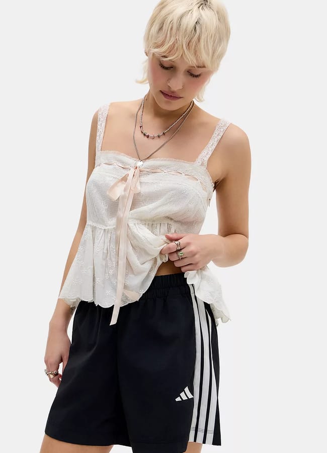 A person with short blond hair wears a white lace top with a bow and black Adidas sports shorts, looking down while holding part of their shirt—showcasing one of the stylish alternatives to denim shorts that are redefining today’s fashion trends.