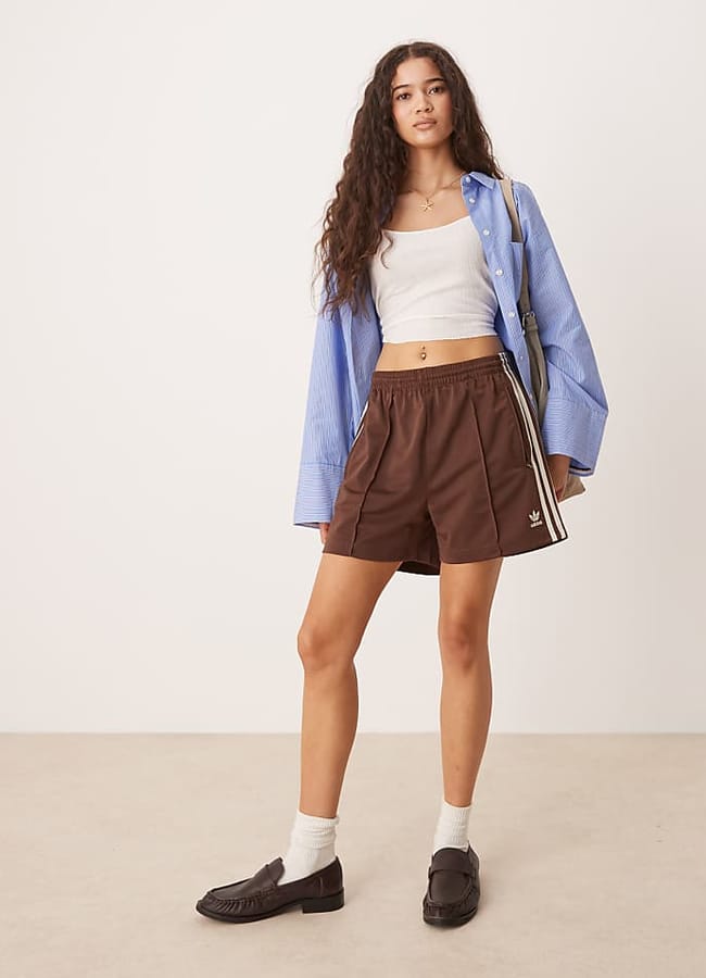 A woman with long curly hair wears a blue shirt over a white crop top, embracing fashion trends with her brown Adidas shorts, white socks, and brown loafers. She stands against a plain background, holding a striped shoulder bag.