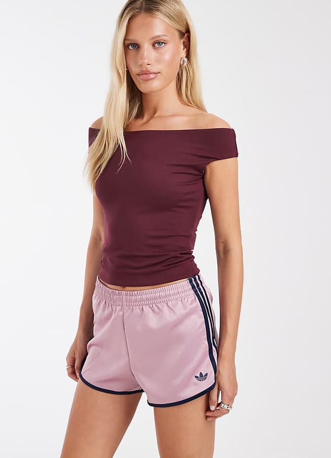 A woman with long blonde hair wears an off-the-shoulder maroon top and light pink Adidas shorts with navy stripes and trim, standing against a plain white background—a stylish example of how denim alternatives are becoming popular in current fashion trends.
