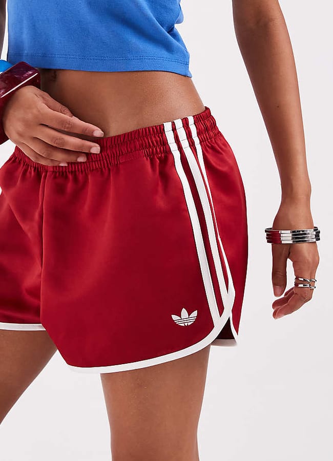 Person wearing red Adidas shorts with white stripes and a blue crop top, showing midriff. Embracing current fashion trends, they accessorise with bracelets on their wrist and rest their hand on their hip. The background is plain white—an outfit that stands out as one of the best denim alternatives this season.