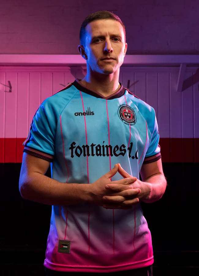 A person stands indoors with hands clasped, wearing a turquoise and pink football core football shirt with Fontaines D.C. on the front and an embroidered crest, under dramatic pink and purple lighting.