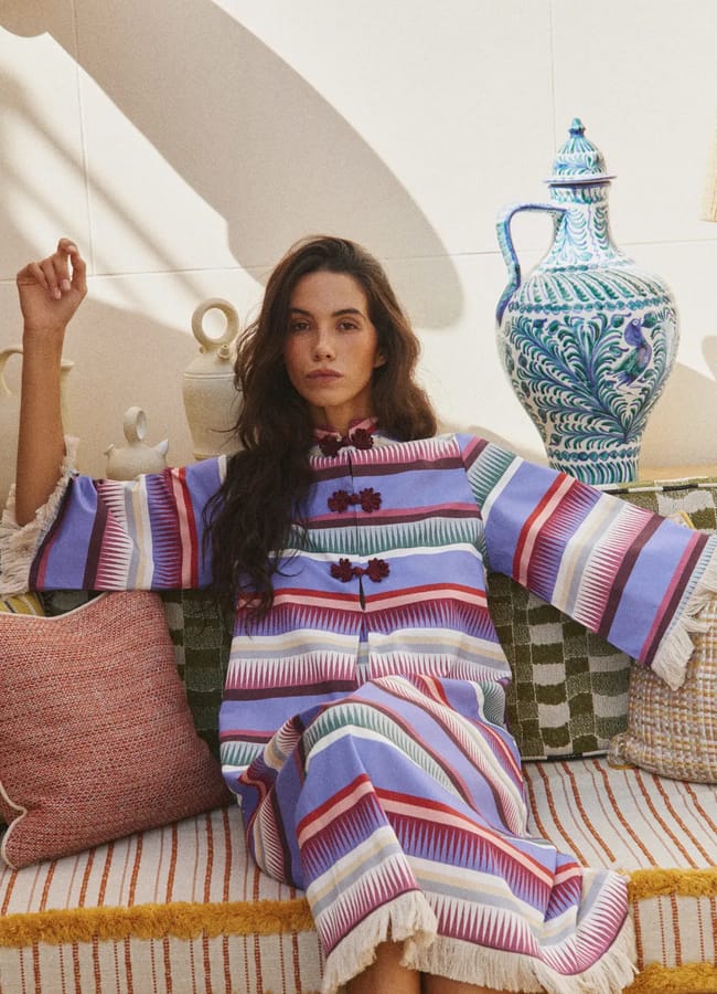 A chic woman in a colourful striped dress sits on a patterned sofa, surrounded by cushions and ceramic vases, with sunlight streaming in from the left.