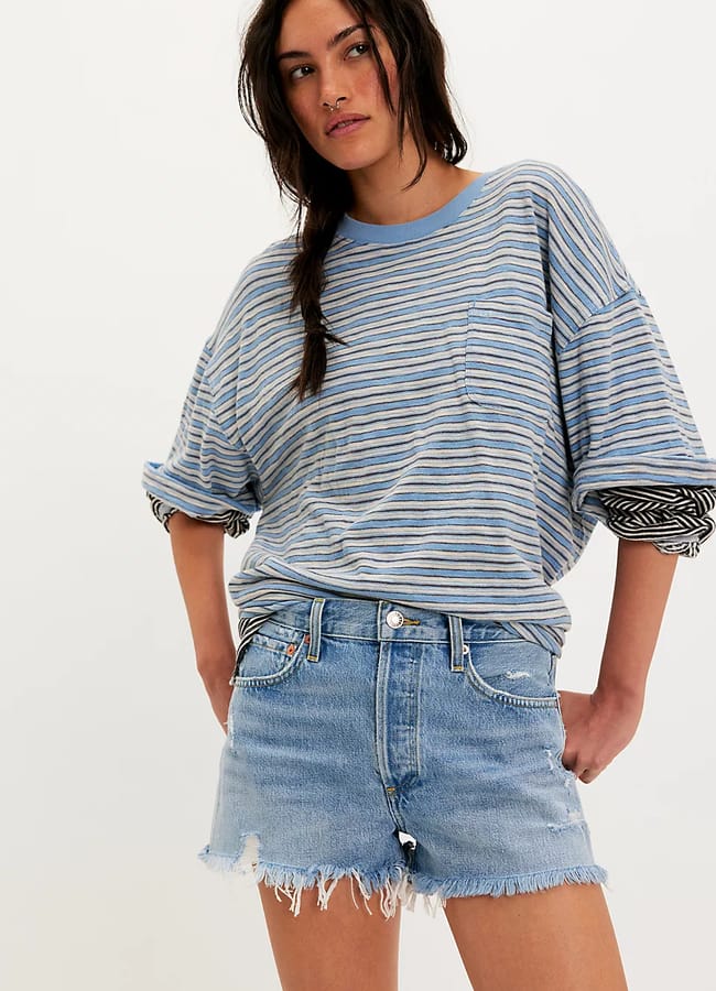 A woman wearing a loose, blue and grey striped T-shirt with rolled sleeves and the best denim shorts—light blue, distressed, and casual—stands with her hands in her pockets against a plain, light background.