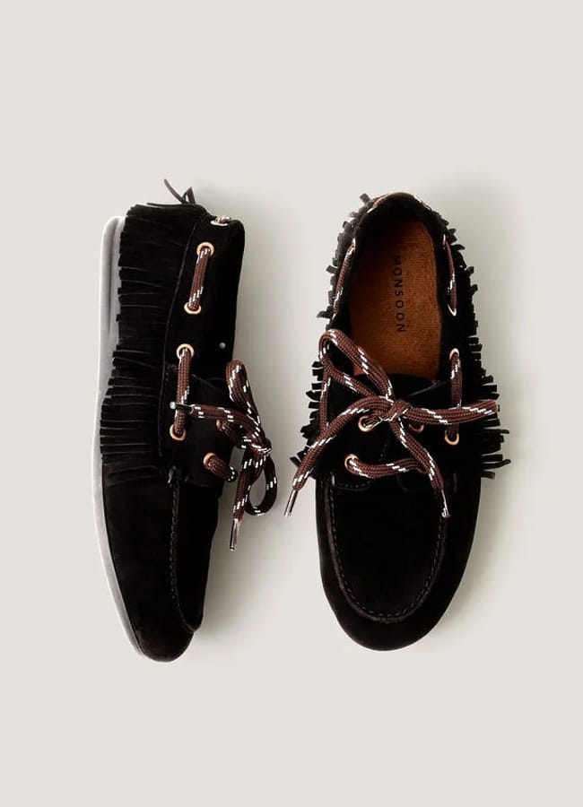 A pair of black suede moccasins with brown leather laces, fringe detailing around the sides, and light brown insoles—perfect for elevating your high street fashion look. One shoe is shown from above and the other from the side.