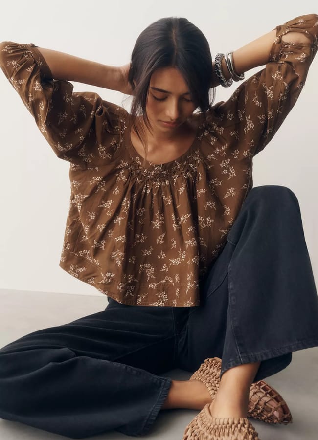 A woman sits on the floor with arms raised, wearing a brown floral blouse, wide-leg dark trousers, woven shoes, and bracelets—a look that perfectly captures high street fashion. She looks down, her dark hair tied back, against a plain background.