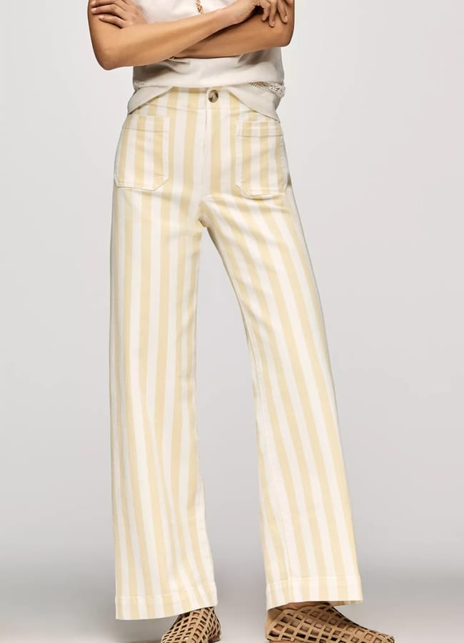 A person wearing high-waisted, wide-leg trousers with vertical yellow and white stripes—a nod to summer micro trends—a tucked-in light top, and woven pointed-toe flats. Their arms are crossed, and their face is not visible.