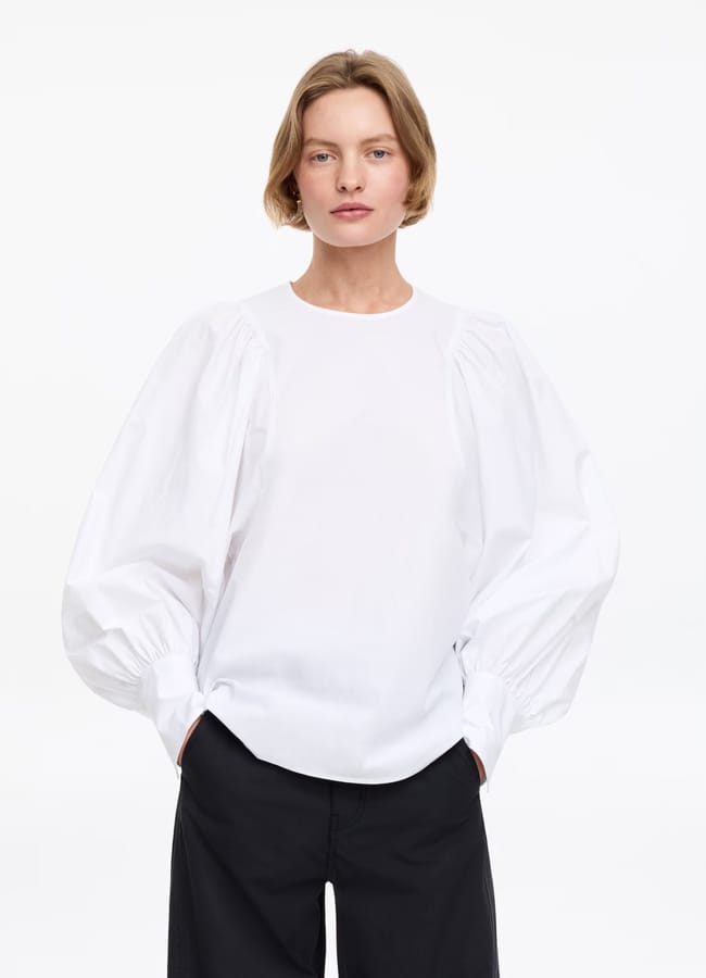 Person with short blonde hair wears a white blouse with voluminous sleeves and black trousers, showcasing a chic high street fashion look. Standing against a plain white background, hands in pockets, looking at the camera.