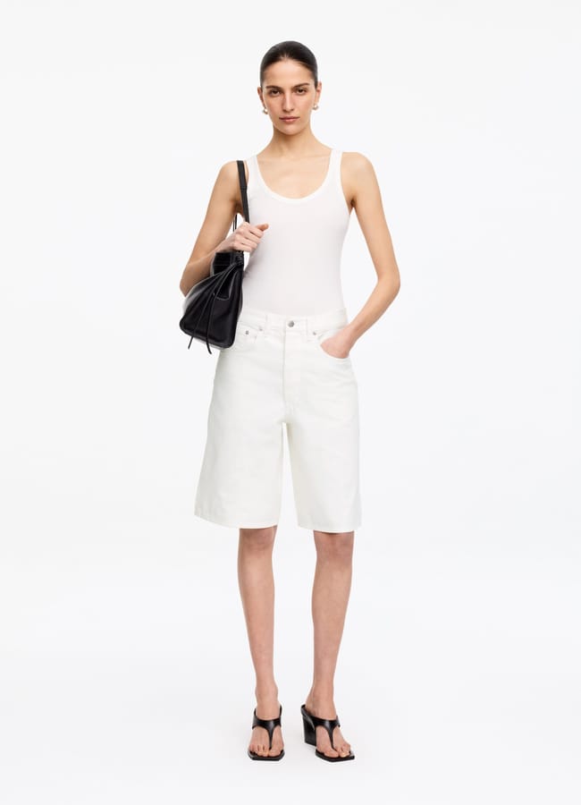 A woman stands against a white background, wearing a white vest top, best denim shorts in knee-length style, black heeled sandals, and carrying a black handbag on her shoulder. She has her hair tied back and a neutral expression.