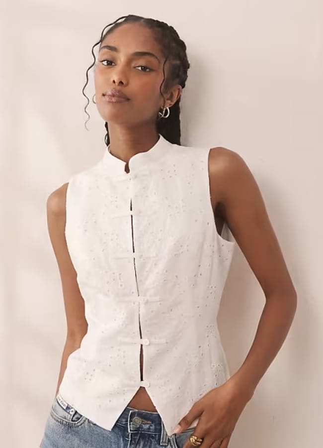 A woman wearing a sleeveless, white button-up top with a mandarin collar and broderie anglaise details stands against a light background, with one hand in her pocket and a calm expression—a subtle style detail ideal for those seeking chic outfit inspiration.