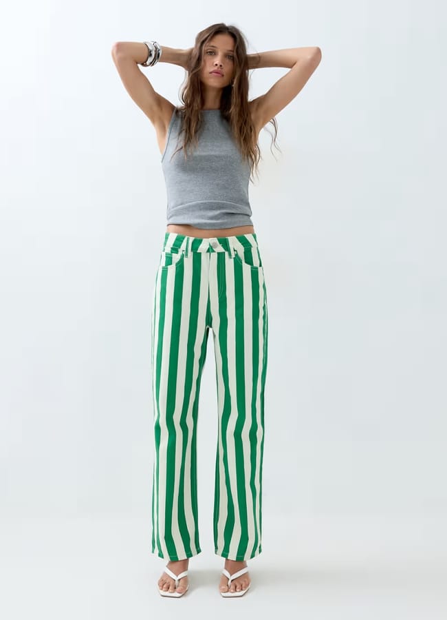A woman stands with her hands on her head, wearing a grey sleeveless top, green and white striped trousers, white sandals, and bracelets—an effortlessly chic nod to summer micro trends against a plain light background.