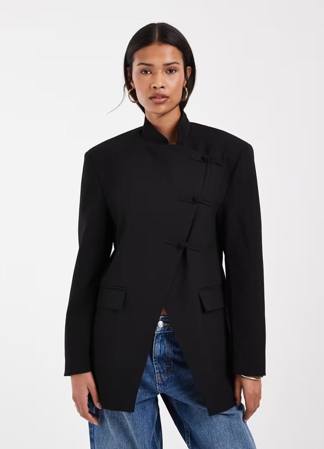 Channelling Kendall Jenner style, a woman wearing a black mandarin-collar blazer with toggle fastenings, paired with blue jeans, stands against a plain white background.