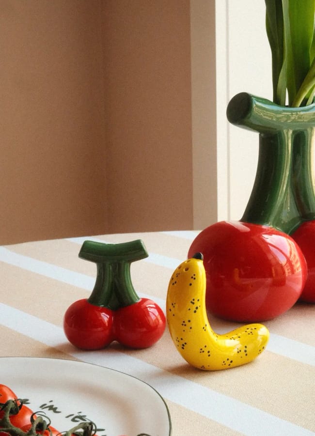 Ceramic cherry and banana sculptures sit on a striped table next to a glass plate with tomatoes, creating a vibrant tableau perfect for the foodie or homewares enthusiast; in the background, a vase with green stems and a red base adds an elegant touch by the window.