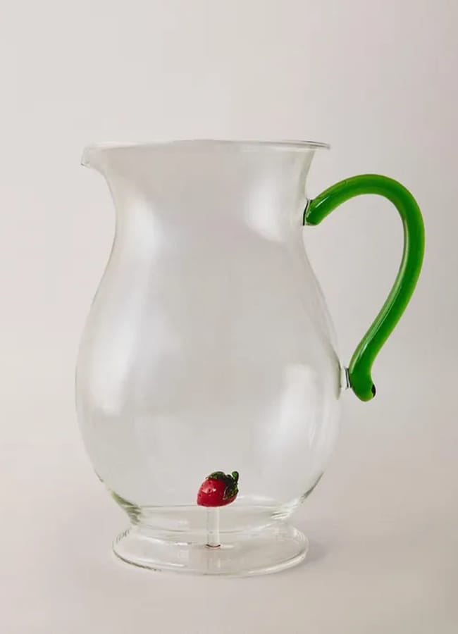 A clear glass jug with a bright green handle and a small red glass strawberry ornament attached to the inside bottom, perfect for adding a touch of charm to your collection of gourmet gifts and unique kitchen accessories.