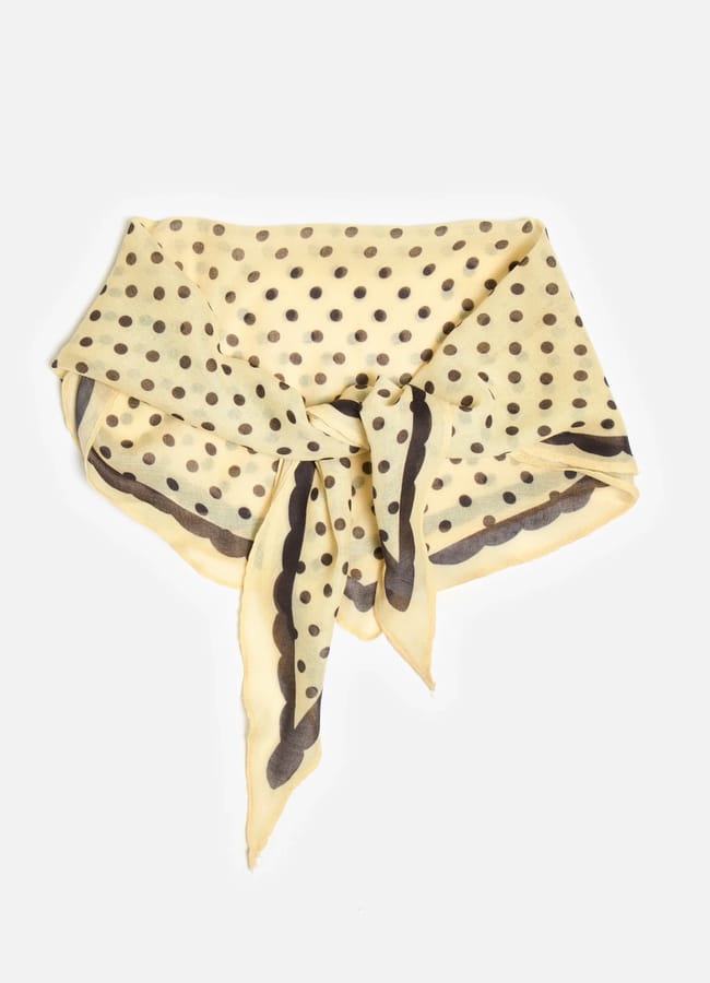 A yellow square scarf with black polka dots and a black border, tied in a simple knot against a white background—an effortlessly chic accessory and the perfect answer to what to wear in Ibiza.