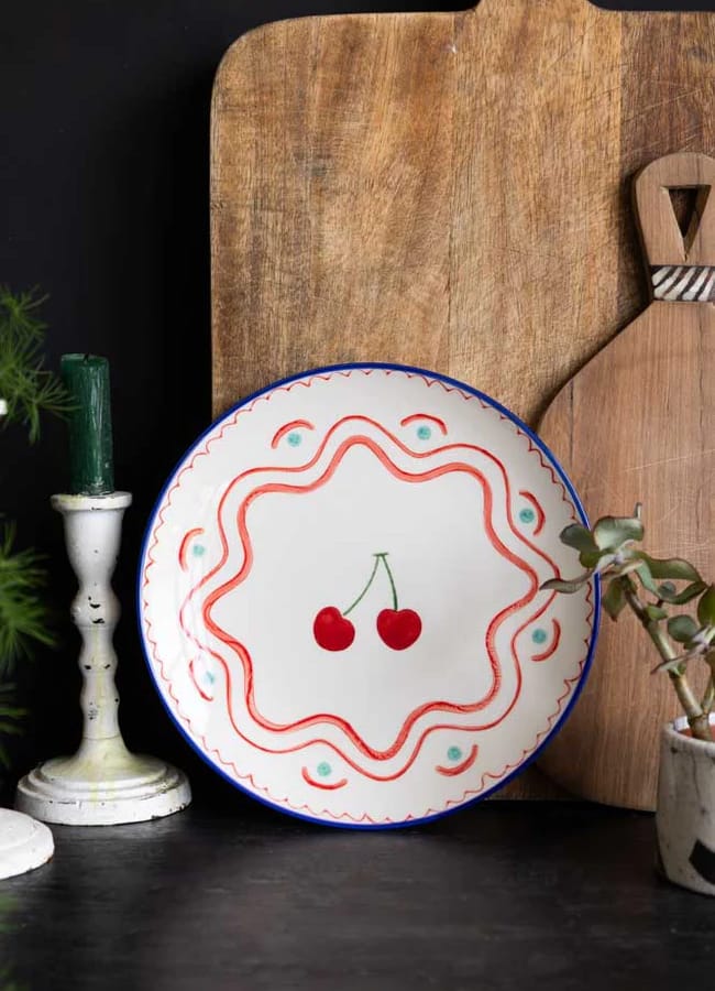 A decorative plate—perfect for any foodie—features two cherries in the centre with red, blue, and green patterns. This charming piece of homeware is placed in front of wooden boards, with a candle and a small potted plant nearby.