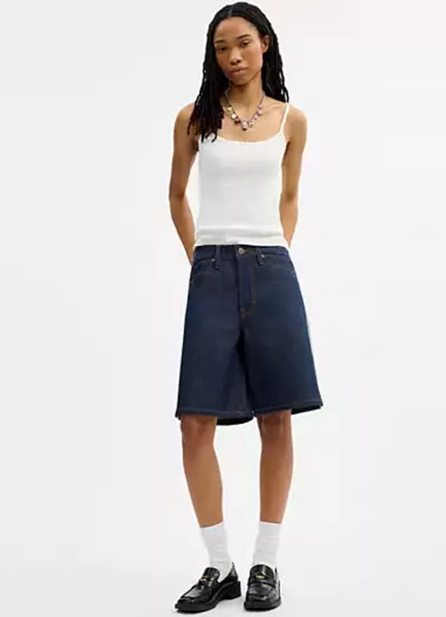 A person wearing a white vest, best denim shorts in a dark knee-length style, white socks, and black shoes stands against a plain white background.