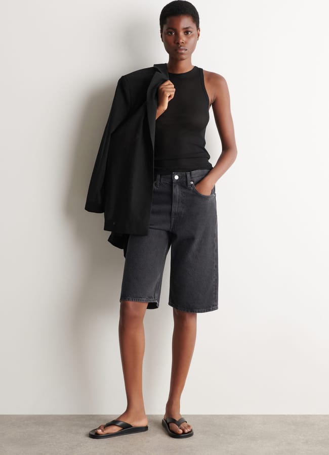A person stands against a white wall, wearing a black sleeveless top, the best denim shorts in black, and black sandals. They hold a black jacket over one shoulder and have one hand in their pocket.