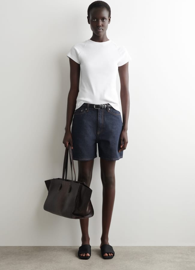 A person stands against a white wall wearing a white T-shirt, the best denim shorts with a belt, black slider sandals, and holding a large black tote bag.