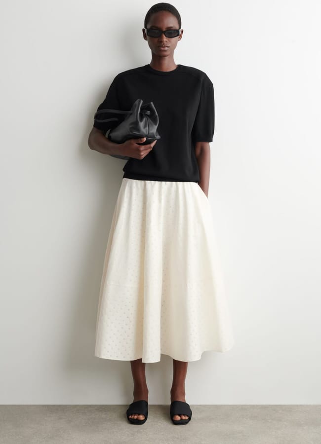 A person wearing sunglasses, a black short-sleeve top, and a white midi skirt stands against a plain wall, showcasing high street fashion while holding a black clutch and wearing black slider sandals.